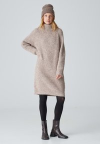 Strickkleid und Beanie in hellbraun mit strukturierter Oberfläche. Das Kleid hat einen hohen Kragen und eine lockere Passform, mit langen Ärmeln und geradem Saum. Schwarze Leggings und Ankle Boots runden den Look ab.