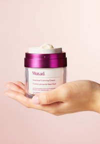 Murad Heartleaf Calming Cream i en genomskinlig burk med ett lila lock, som visar en krämfärgad produkt med en mjuk textur, hålls i en hand.