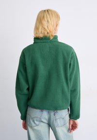 Pull en polaire vert avec col montant et tissu texturé, associé à un jean en denim bleu clair. Design simple, sans matériel visible.