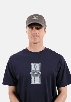 Homme portant une casquette grise à logo floral et un t-shirt bleu marine avec le texte vertical OXBOW et un motif ovale au centre sur fond blanc.