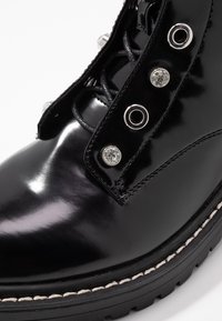 Černé lakované kožené kotníkové boty s hladkým povrchem, dekorativními stříbrnými akcenty a silnou podrážkou s viditelným šitím.