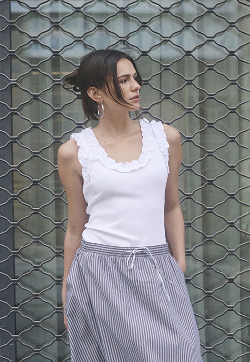 Top blanco con escote con volantes combinado con una falda a rayas con cintura ajustable. La modelo se encuentra de pie frente a un fondo de rejilla metálica.