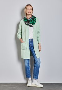 Hellgrüner langer Cardigan mit zwei Fronttaschen, getragen über einem weißen T-Shirt und blauen Jeans. Ein mehrfarbiger gestreifter Schal sorgt für Textur.