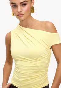 Haut asymétrique jaune avec des détails drapés, en tissu doux, à manches courtes, texture froncée, complété par de grandes boucles d'oreilles dorées.