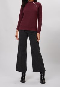 Pull côtelé bordeaux à manches longues, doté d'un col montant et de trois boutons argentés sur l'épaule gauche, associé à un jean noir à jambes larges.