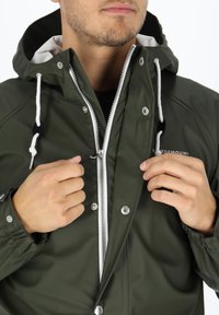 Donkergroene waterdichte jas met een ritssluiting aan de voorkant, contrasterende witte voering en verstelbare trekkoordjes aan de capuchon en manchetten. Zilverkleurige drukknoopjes zichtbaar.