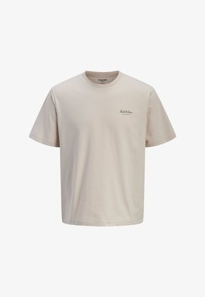 Boy standing wearing a light beige T-shirt, wide faded brown pants, and white sneakers against a plain light background.