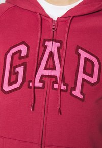 Persona che indossa una felpa rossa con zip e grandi lettere rosa "G.A.P" ricamate sul davanti.