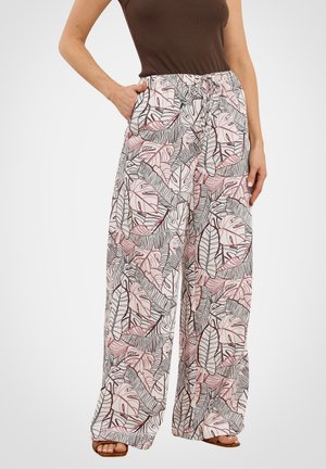 Frau trägt weite Hose mit schwarzen und pinken tropischen Blattmustern und braunem ärmellosen Oberteil, steht mit einer Hand in der Tasche.