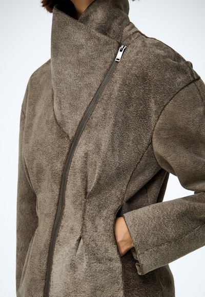 Barna műszőrme kabát magas gallérral, oldalsó cipzárral és elülső zsebekkel. Texturált felület és lazább szabás.