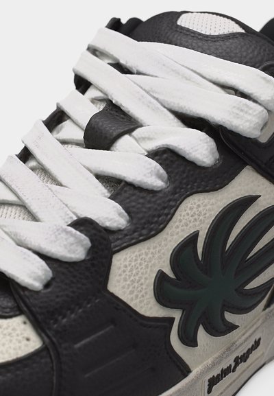 Ασπρομαύρο αθλητικό παπούτσι με λευκά κορδόνια και σκούρο πράσινο στυλιζαρισμένο φύλλο λογότυπο στο πλάι.