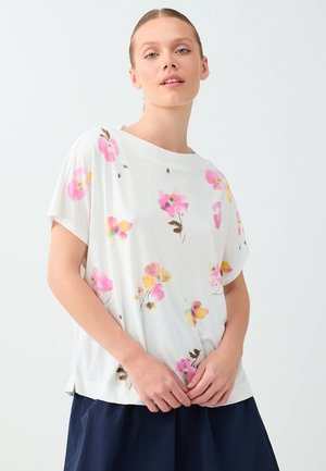 Młoda kobieta w białej koszulce z krótkim rękawem ozdobionej różowo-żółtymi kwiatowymi wzorami oraz w ciemnej spódnicy, stojąca na jednolitym tle.