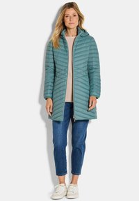 Teal pufferjas met capuchon en ritssluiting aan de voorkant; gekenmerkt door een chevron-gestikt patroon, lange mouwen en valt onder de knieën. Gecombineerd met blauwe spijkerbroeken.