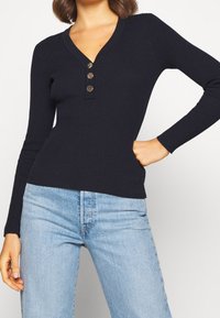 Marineblauw geribd shirt met lange mouwen, een V-hals en drie decoratieve knopen bij de halslijn, gecombineerd met lichtblauwe spijkerbroek. Soepele textuur.