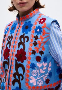 Γυναίκα που φοράει μπλε γιλέκο κεντημένο με πολύχρωμα λουλούδια και πορτοκαλί μοτίβα από κληματόβεργες πάνω από ανοιχτό μπλε πουκάμισο με ρίγες.