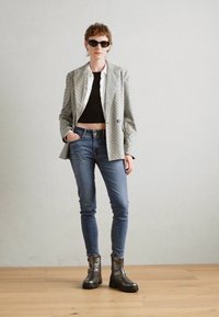 Siv štrihan blazer čez črn kratek top, modre ozke kavbojke in črne masivne škornje. Model nosi temna sončna očala. Enostavna kulisa.