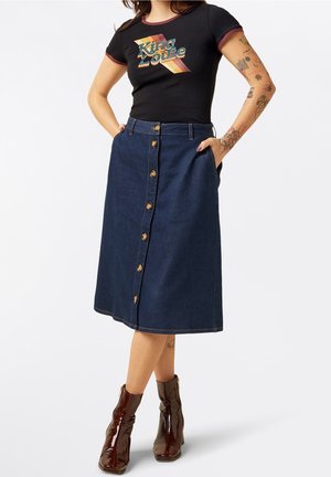 Frau trägt schwarzes T-Shirt mit Grafik, dunklen, knielangen Jeansrock mit Knöpfen und glänzende braune Stiefeletten, mit tätowierten Armen und Händen in den Taschen.