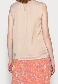 Ærmeløs beige bluse med en blød, glat tekstur. Har en nøglehulåbning bagpå og er parret med en blomsterprintet pink nederdel.