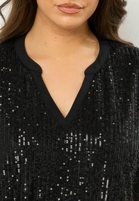 Schwarzes, mit Pailletten besetztes Oberteil mit V-Ausschnitt und glattem schwarzen Besatz. Die Pailletten erzeugen einen schimmernden Effekt auf dem strukturierten Stoff.