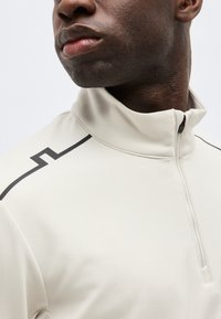 Κοντινό πλάνο ενός άνδρα που φοράει μπεζ πουλόβερ με τετραπλό φερμουάρ και μαύρη γωνιακή ρίγα στον ώμο, με φόντο απλό.