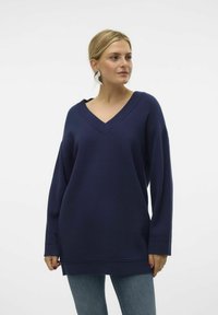 Marineblauer, oversize V-Ausschnitt-Pullover aus weichem Stoff mit langen Ärmeln und leicht abgerundetem Saum, kombiniert mit Jeans aus Denim.
