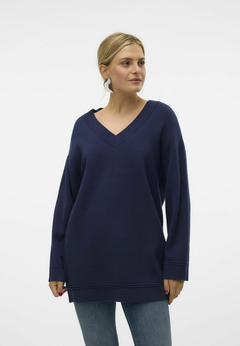 Marineblauer, oversize V-Ausschnitt-Pullover aus weichem Stoff mit langen Ärmeln und leicht abgerundetem Saum, kombiniert mit Jeans aus Denim.