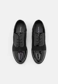 Para czarnych lakierowanych i matowych skórzanych butów oxford z sznurówkami, widziana z góry na jednolitym białym tle.