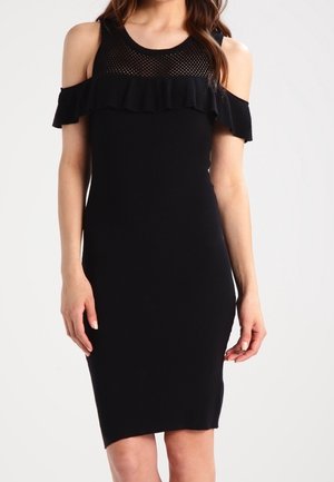 Robe noire à épaules dénudées avec détail volanté et empiècement en résille au niveau de l'encolure, portée par une femme debout.