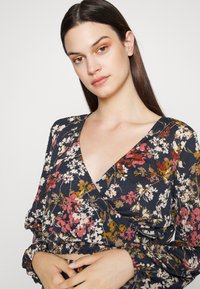 Top envolvente floral com um decote em V profundo, pequenas mangas bufantes e um fundo azul-marinho escuro com flores em tons de rosa, branco e mostarda.