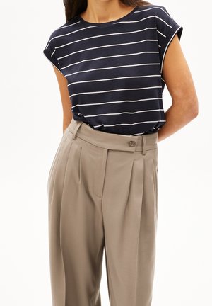 Nainen, jolla on päällään tummansininen lyhythihainen yläosa, jossa on valkoiset vaakaraidat, ja joka on sullottu korkeavyötäröisten, beigejen, laskostettujen housujen sisään, joissa on nappikiinnitys.