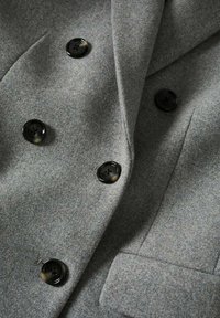 Grå uldblandings dobbeltknapsfrakke med seks store sorte knapper. Har en klappet lapel og en tekstureret finish med et poleret udseende.