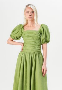 Robe en lin vert avec des manches bouffantes, décolleté carré et détails froncés à la taille, se déclinant en une jupe ample.