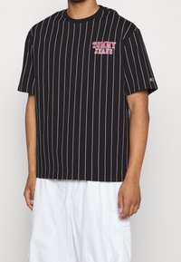 Muž v černém tričku s vertikálním pruhovaným vzorem a logem "Tommy Jeans" na hrudi, v bílé volné kalhoty, stojící proti bílému pozadí.