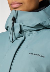Detailný záber osoby so svetlomodrou vodeodolnou bundou Didriksons s vysokým golierom a čiernym zipsom.