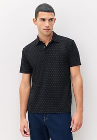 Joven vestido con una camiseta polo negra de manga corta con pequeños patrones de cruces doradas, combinada con unos jeans azul oscuro, de pie contra una pared blanca.