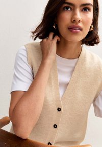 Giovane donna con capelli corti castano che indossa orecchini a cerchio dorati, una maglietta bianca e un gilet beige con bottoni, seduta e mentre si tocca i capelli.