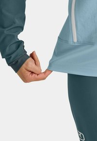 Light blue athletic top with a front zipper, long sleeves, and thumbhole cuffs. The hand is pulling at the bottom hem, revealing a darker base layer.
