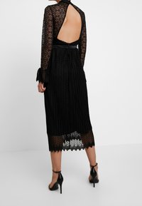 Robe en dentelle noire à manches longues, dos ouvert, texture plissée et ourlet en forme d'ondulations, associée à des talons hauts noirs à brides.
