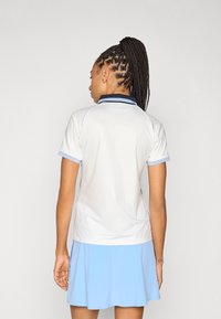 Balta īsroku polo krekls ar tumši zilu apkakli un gaiši ziliem akcentiem, darināts no teksturētas auduma un pieguloša griezuma. Valkāts kopā ar zilu svārku.