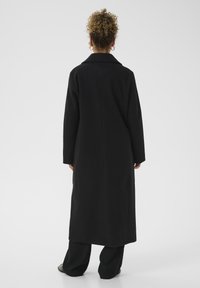 Person with curly hair wearing a long black coat, black pants, and black shoes standing facing away against a plain background.