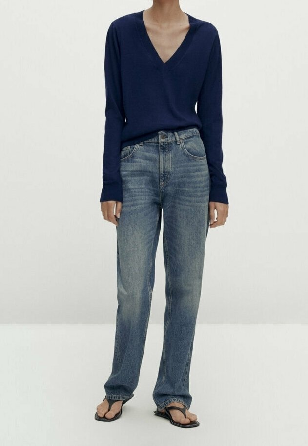 Personne portant un haut à manches longues bleu marine avec col en V, un jean bleu clair droit et des tongs noires, debout devant un fond uni.
