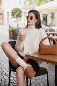 Žena s okuliarmi na slnko a bielym svetríkom sedí pri stolíku v vonkajšej kaviarni, drží ľadovú kávu, na stole je hnedá kabelka.
