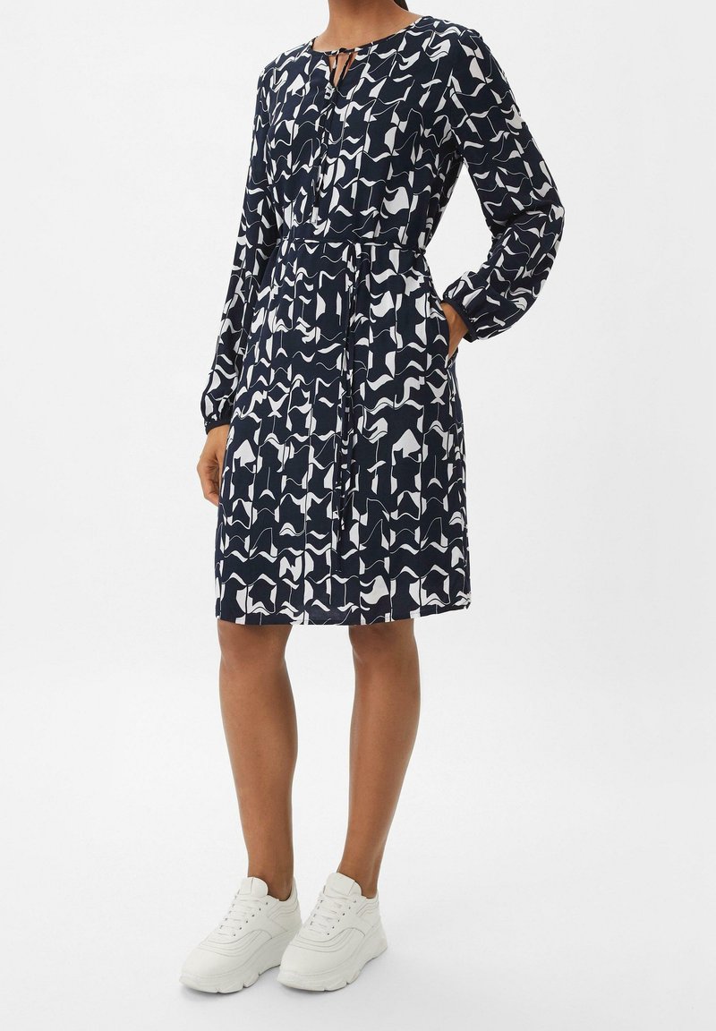 Femme portant une robe à manches longues jusqu'aux genoux, de couleur bleu marine avec un motif blanc, et des baskets blanches, se tenant debout avec une main dans la poche.