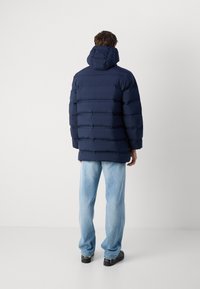 Námornícka prešívaná bunda s kapucňou, ktorá má vodorovné prešívané časti a mierne oversized strih. Nosí sa s svetlomodrými džínsami a čiernymi topánkami.