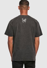 T-shirt de algodón negro con corte relajado, que presenta un gráfico en la parte superior de la espalda con una mariposa blanca y flores. Mangas cortas y costuras visibles.