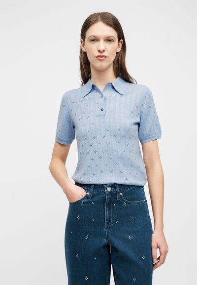 Femme portant un polo en maille à manches courtes bleu clair avec de petits détails ajourés et un jean taille haute bleu foncé avec des motifs brodés subtils.