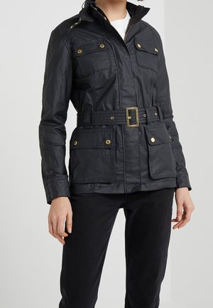 Persoon draagt een zwarte jas met ceintuur, vier klepzakken aan de voorkant en gouden knoopdetails, gecombineerd met een zwarte broek.