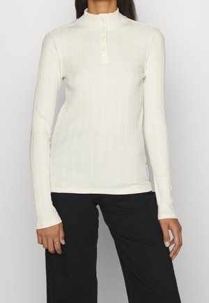 Haut blanc à manches longues côtelé avec un col montant et un plastron à quatre boutons. Présente des manches ajustées et une finition douce et texturée.