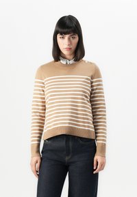 Beiger Strickpullover mit weißen Streifen, Rundhalsausschnitt und drei Knöpfen an der Schulter. Kombiniert mit dunkelblauen Jeans.