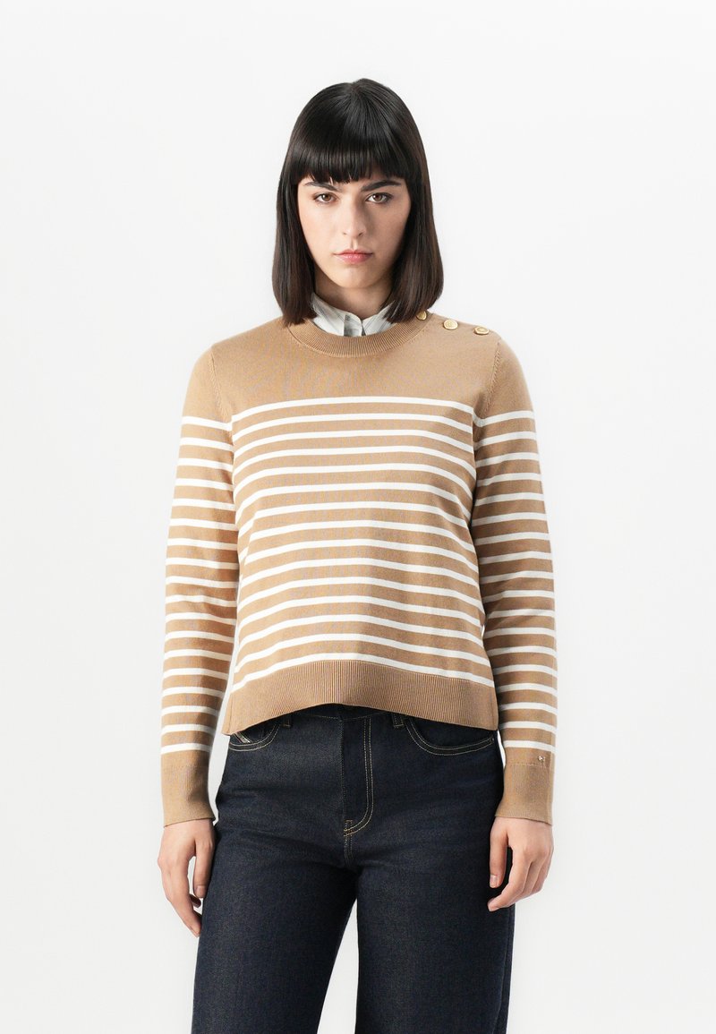 Beiger Strickpullover mit weißen Streifen, Rundhalsausschnitt und drei Knöpfen an der Schulter. Kombiniert mit dunkelblauen Jeans.